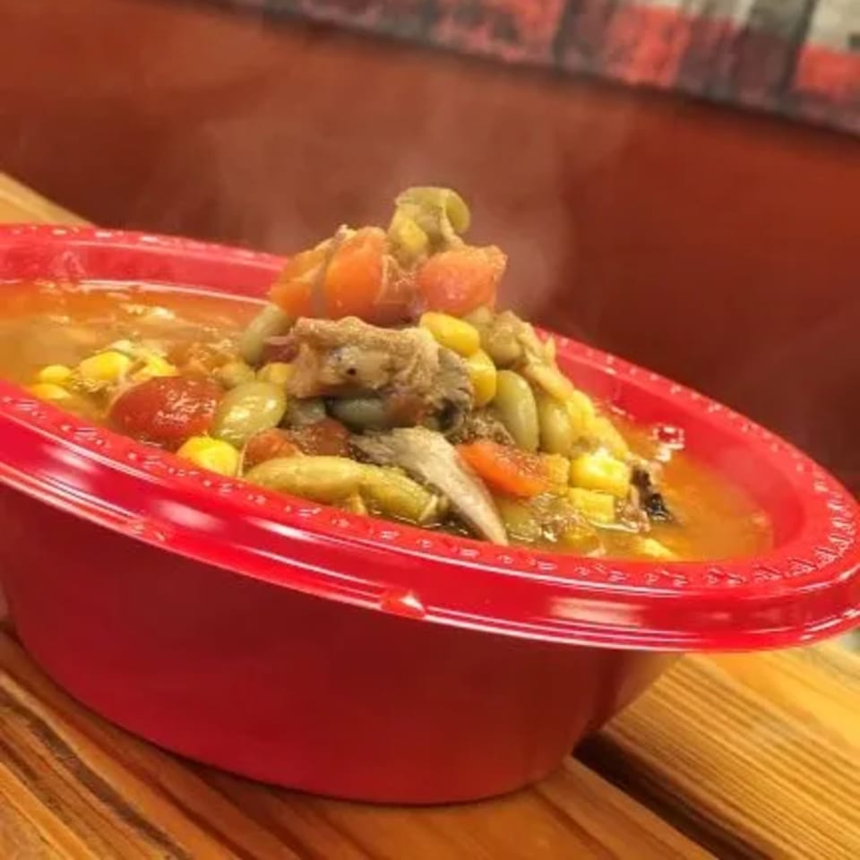 Steaming Brunswick stew with chicken, corn, and tomatoes in a red bowl on a wooden table.