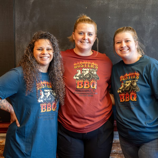 Three smiling women in Buster's BBQ t-shirts stand together against a dark wall.