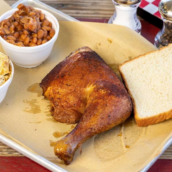 A BBQ chicken leg quarter, baked beans, and white bread on a tray with parchment paper.