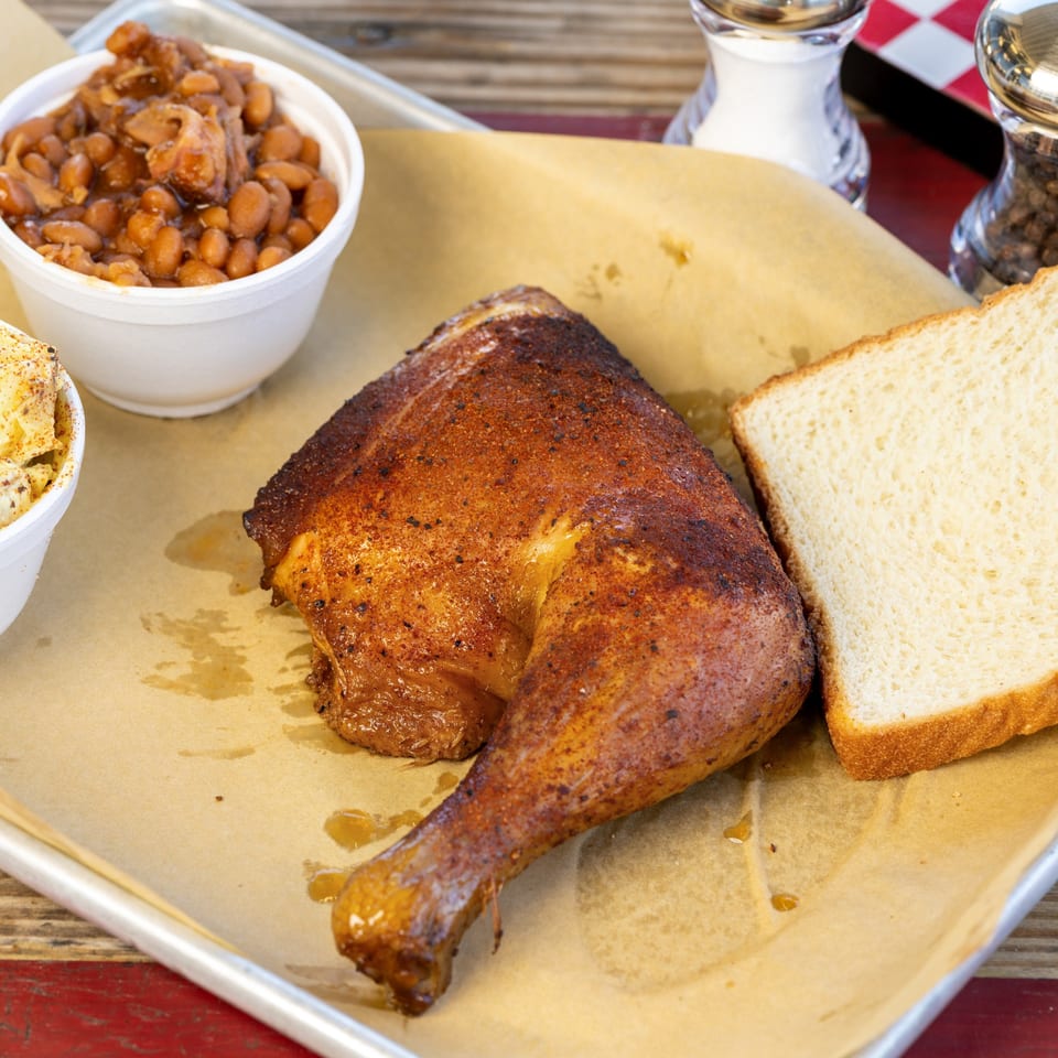 A BBQ chicken leg quarter, baked beans, and white bread on a tray with parchment paper.