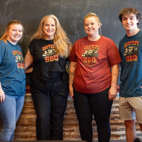Four smiling Buster's BBQ staff members in branded t-shirts stand against a dark wall.