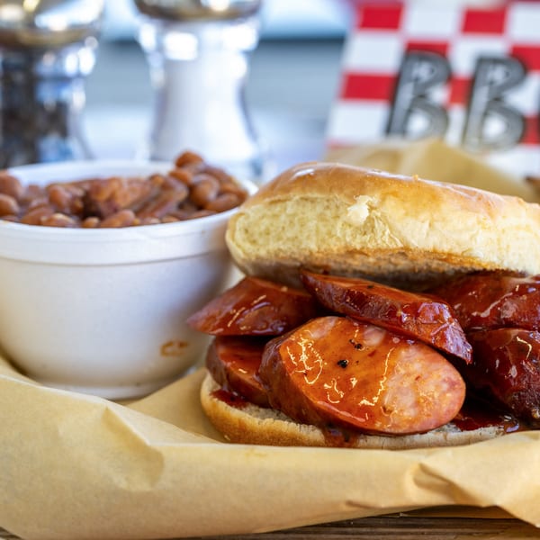 Sausage sandwich with barbecue sauce and a side of baked beans in a white cup.