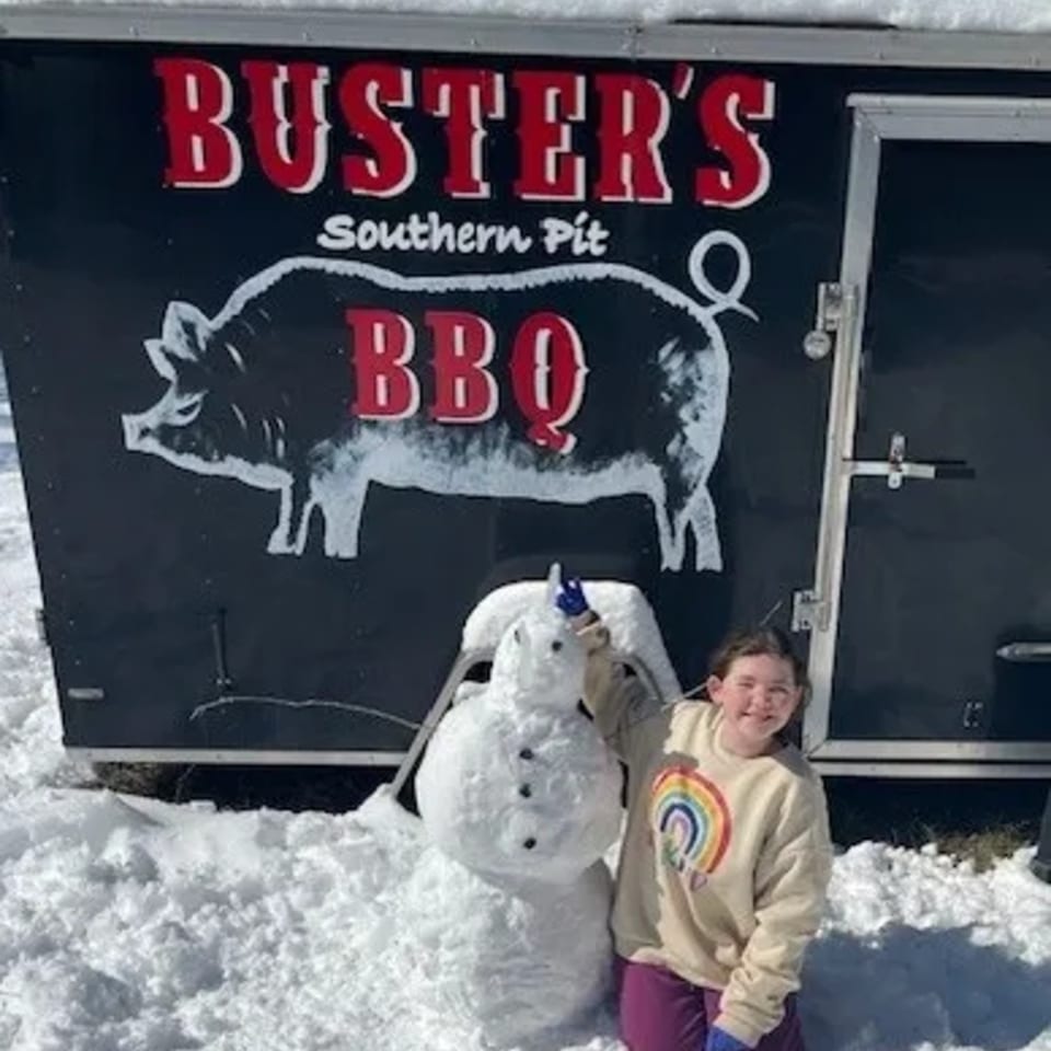 A girl and a snowman stand in front of the Buster's Southern Pit BBQ food truck in the snow.