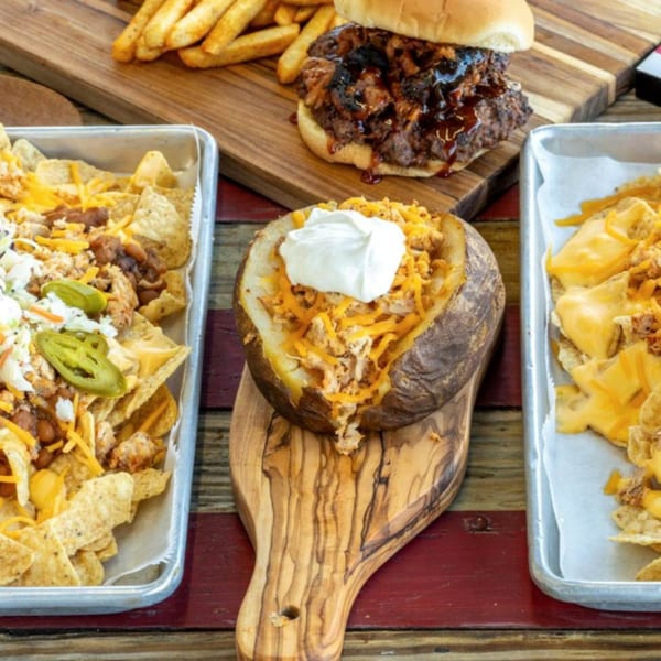 A BBQ burger with fries, two trays of nachos, and a loaded baked potato on a wooden table.
