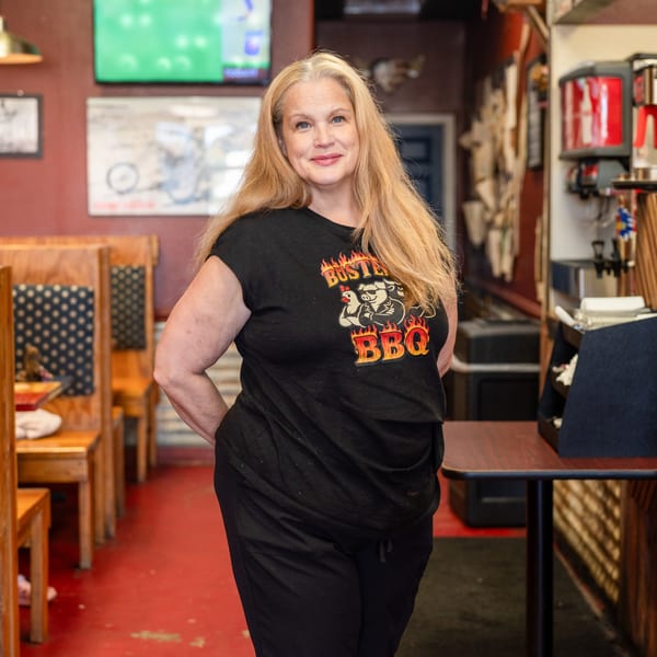 A smiling woman with long blonde hair wearing a black "Buster's BBQ" t-shirt stands in a restaurant.