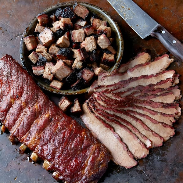 A full rack of BBQ ribs, sliced brisket, and burnt ends on a dark surface with a knife.