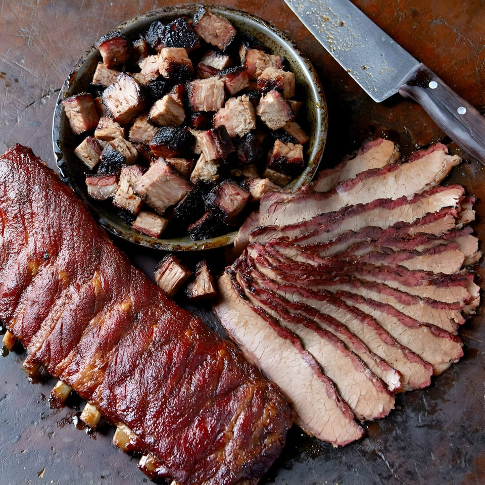 A full rack of BBQ ribs, sliced brisket, and burnt ends on a dark surface with a knife.