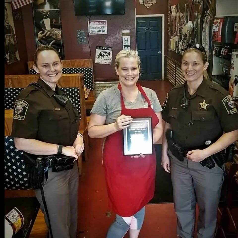 Two female Alabama Sheriff deputies flank a woman in a red apron holding a framed award at Busters BBQ.