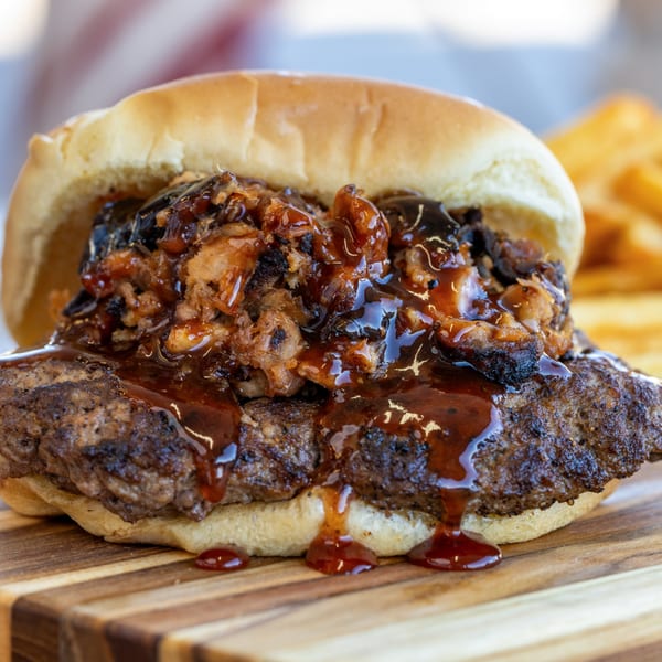Close-up of a juicy burger with barbecue sauce and pulled pork on a bun, with fries in the background.